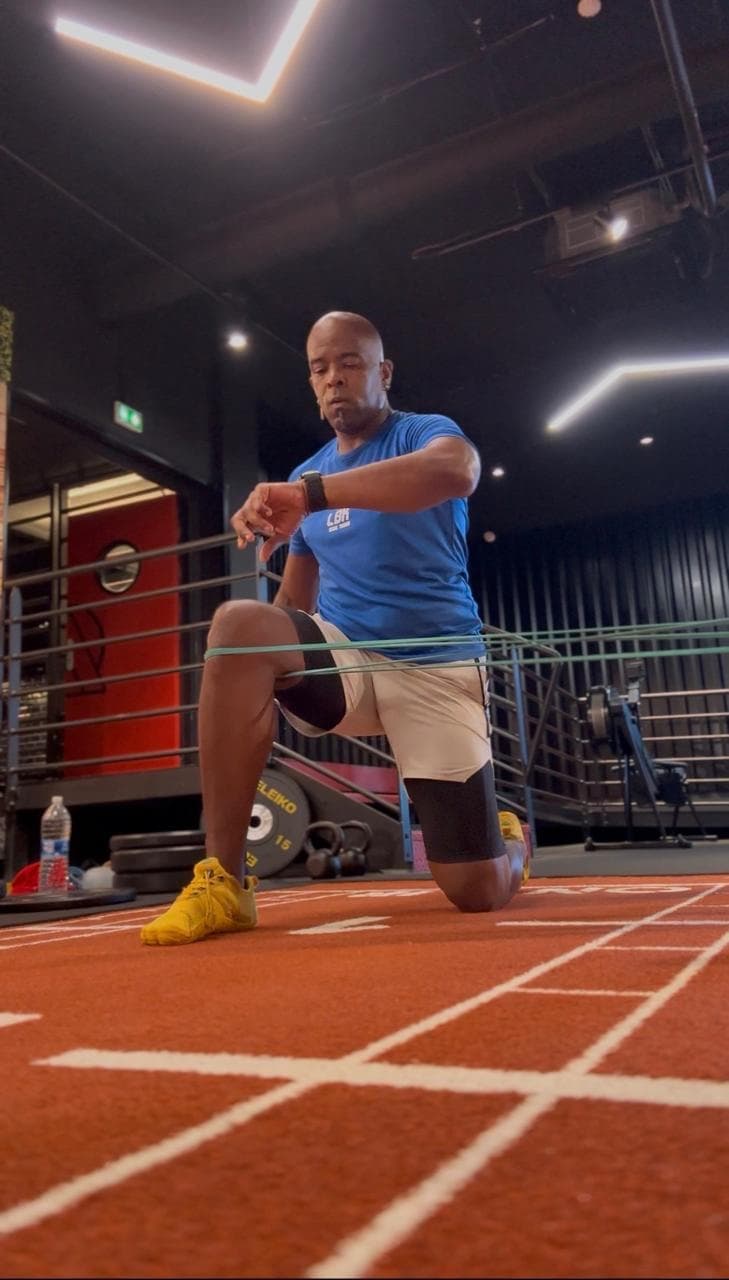 Laurent LABECA en fente au sol, travaillant avec un élastique sur une piste d’athlétisme en salle